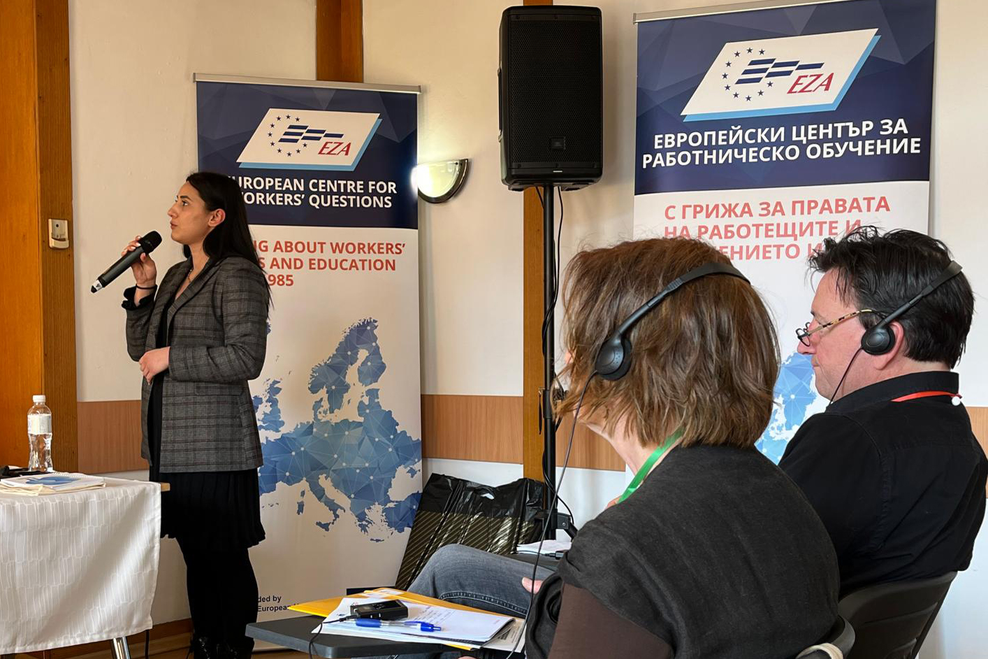 Eine Frau am Mikro spricht, Menschen hören ihr zu.