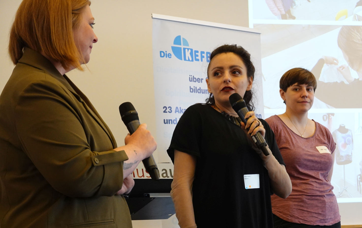 Drei Frauen stehen auf einer Bühne, zwei haben ein Mikrofon in der Hand.