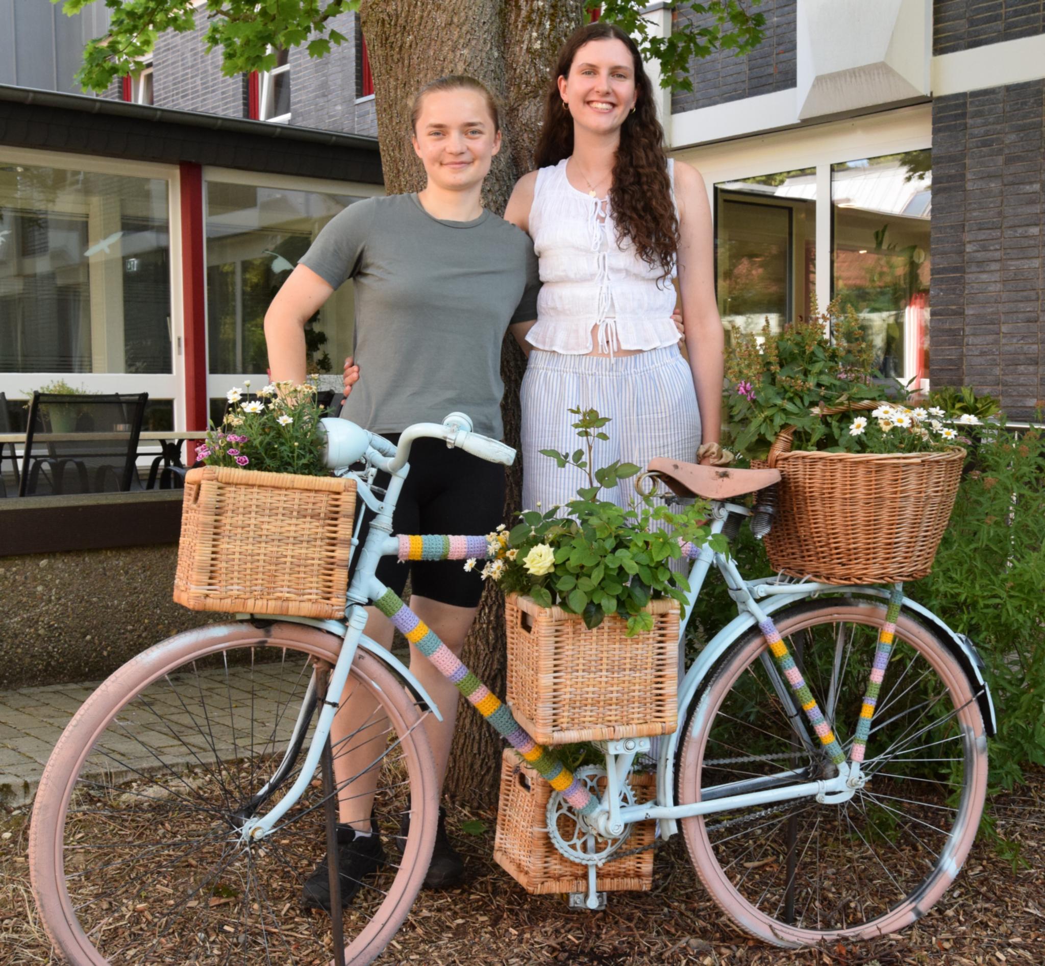 Zwei junge Frauen stehen hinter einem Fahrrad vor einem Baum. Das Fahrrad ist mit Blumen und farbiger Wolle verziert.