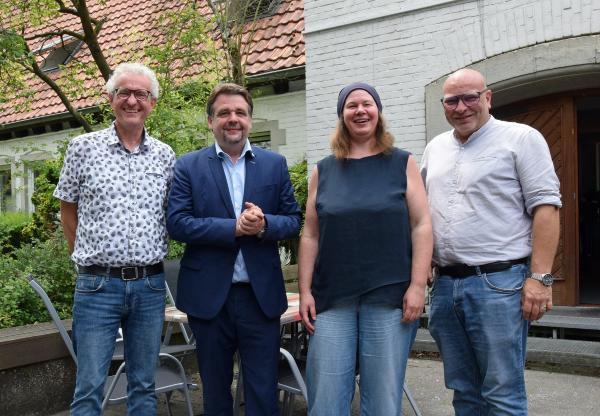Drei Männer und eine Frau stehen in einem Innenhof vor einem Tor und einem Baum.