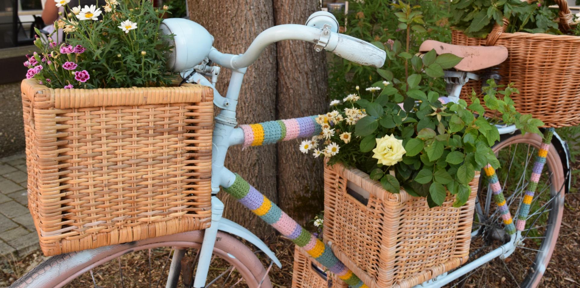 Ein Fahrrad ist mit bunter Wolle, Blumenkästen und Blumen verziert und lehnt an einen Baum.