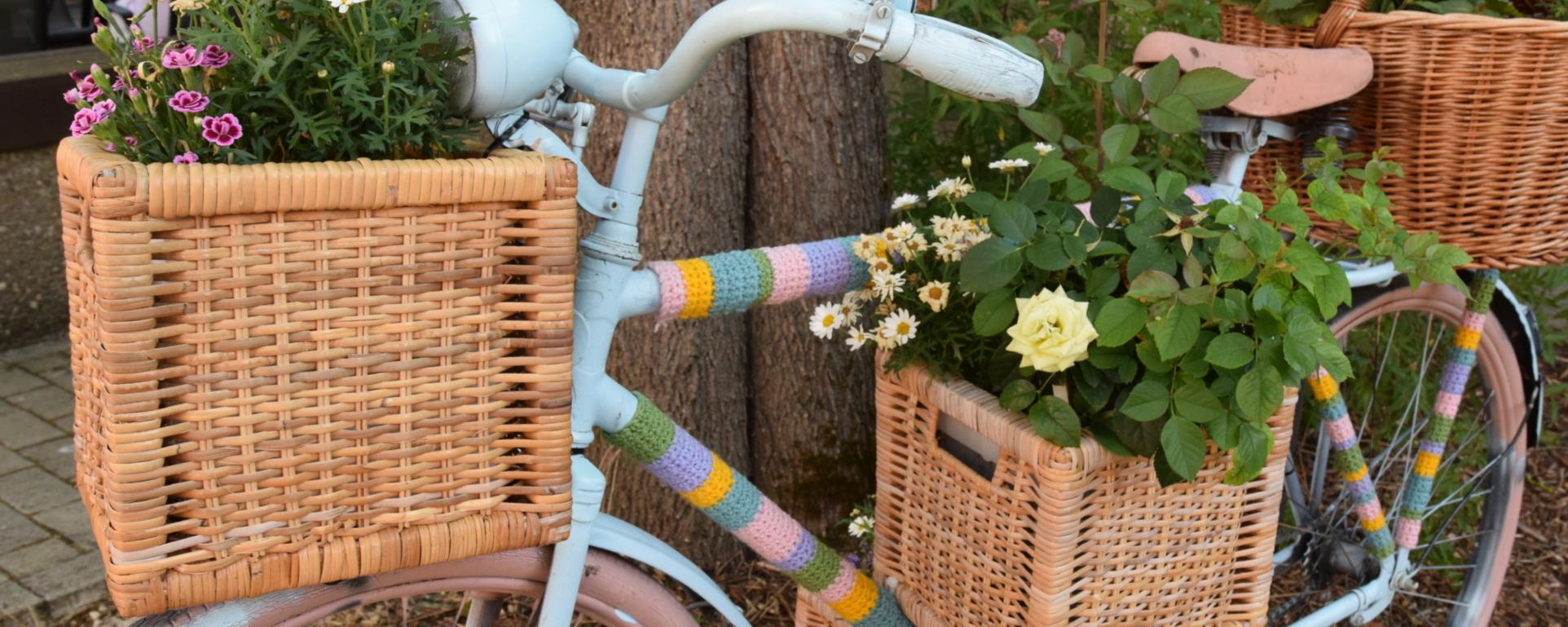 Ein Fahrrad ist mit bunter Wolle, Blumenkästen und Blumen verziert und lehnt an einen Baum.