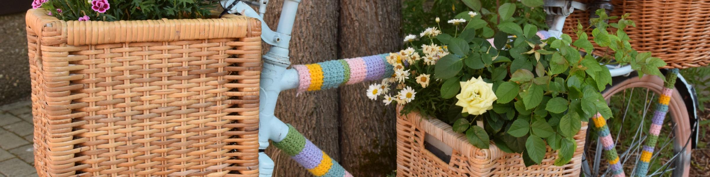 Ein Fahrrad ist mit bunter Wolle, Blumenkästen und Blumen verziert und lehnt an einen Baum.