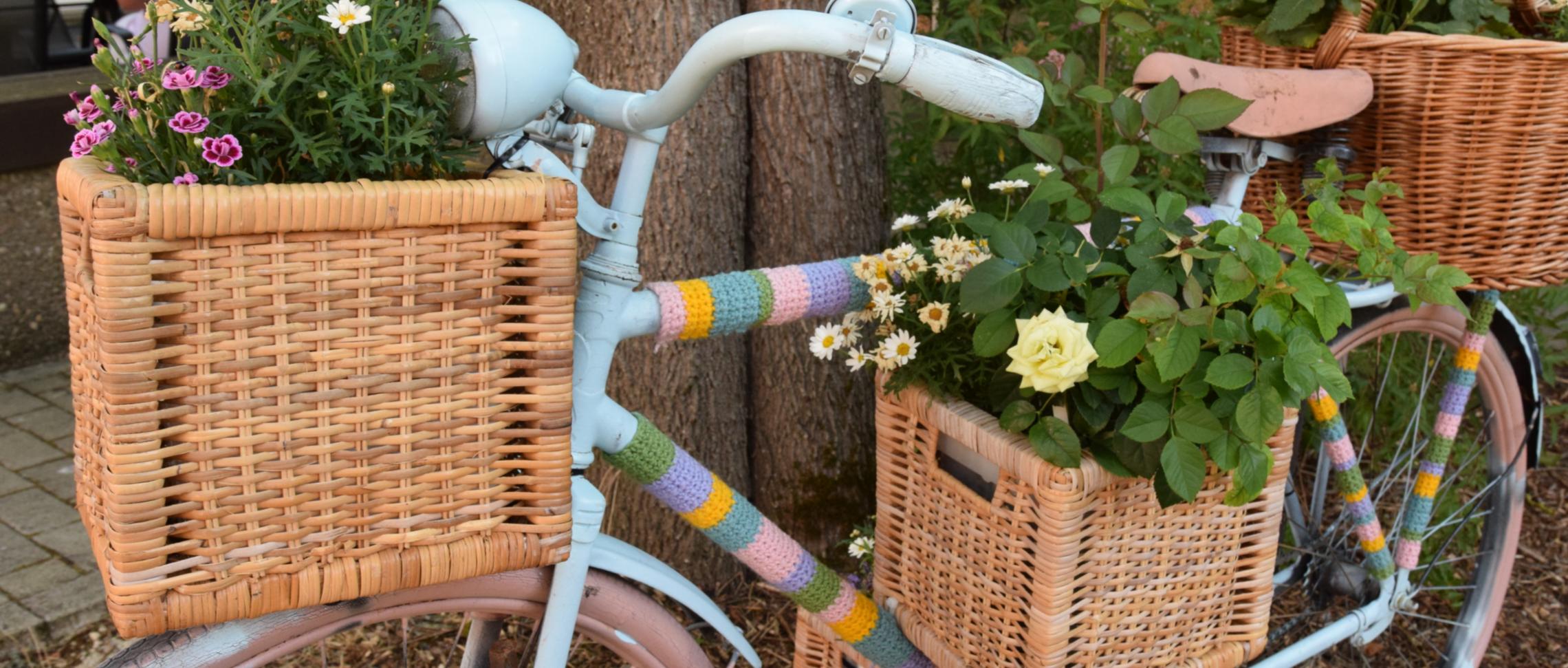 Ein Fahrrad ist mit bunter Wolle, Blumenkästen und Blumen verziert und lehnt an einen Baum.