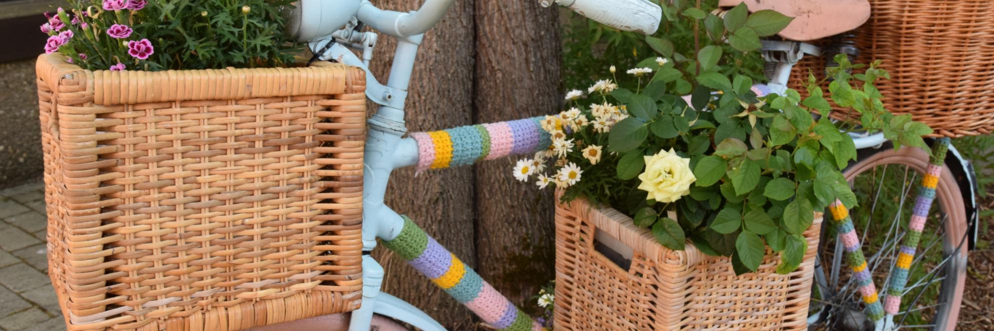 Ein Fahrrad ist mit bunter Wolle, Blumenkästen und Blumen verziert und lehnt an einen Baum.