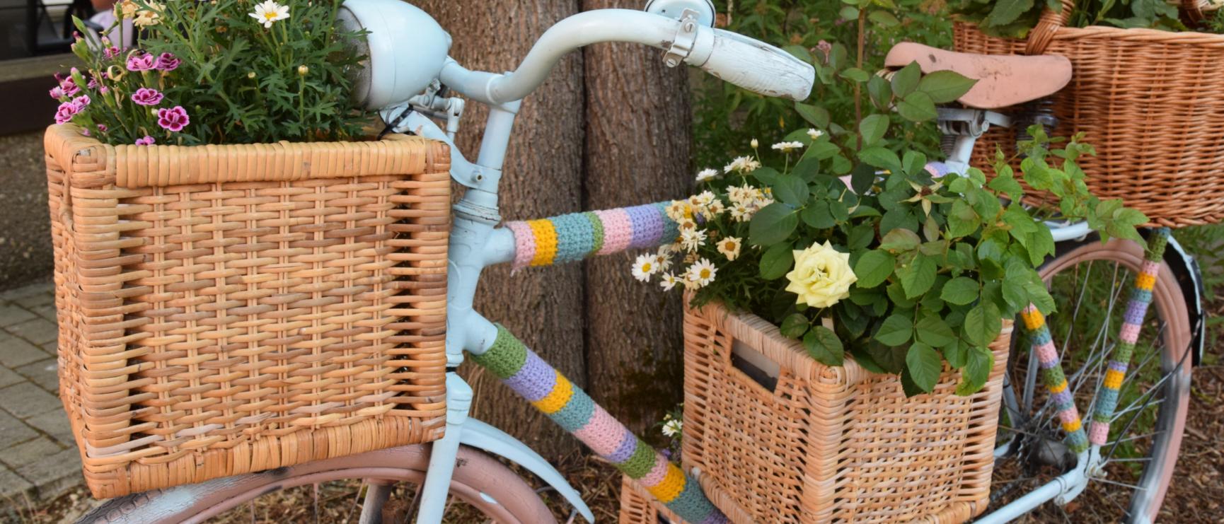 Ein Fahrrad ist mit bunter Wolle, Blumenkästen und Blumen verziert und lehnt an einen Baum.
