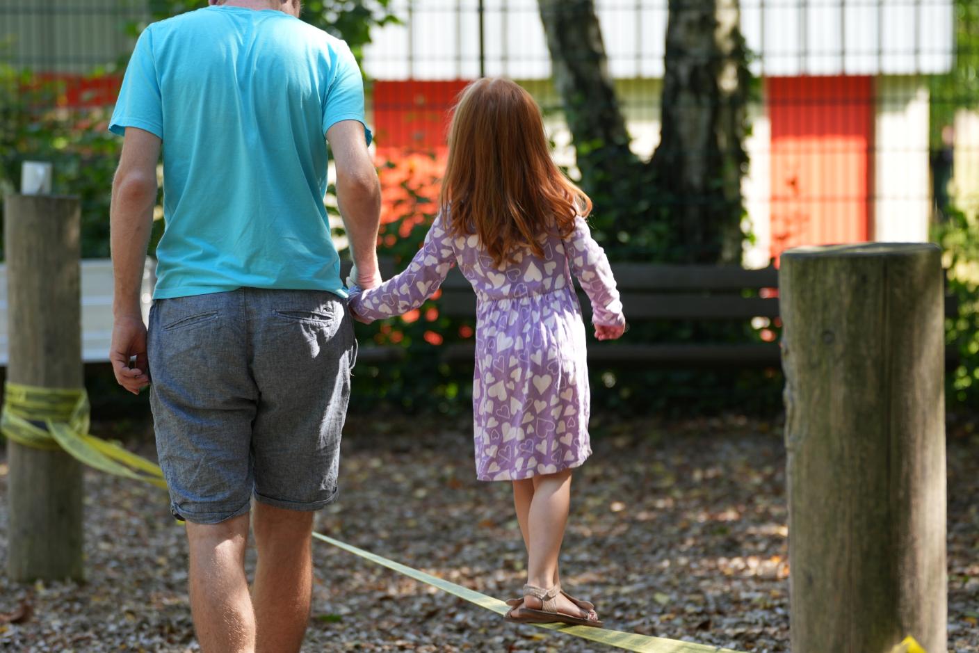 Ein Kind balanciert auf einem Niedrigseil. Ein Mann hält sie an der Hand.