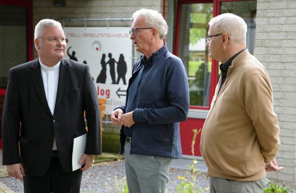 Drei Männer stehen zusammen. Im Hintergrund ein Plakat, auf dem Nell-Breuning-Haus steht.