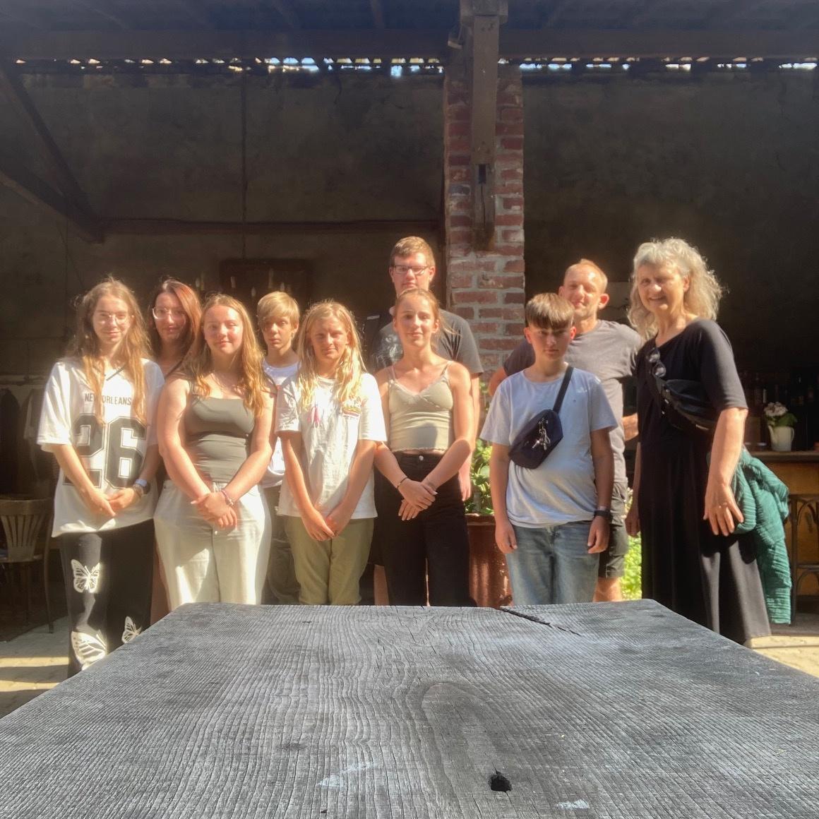 Gruppe von neun Personen steht vor einer Backsteinwand unter einem überdachten Bereich an einem sonnigen Tag